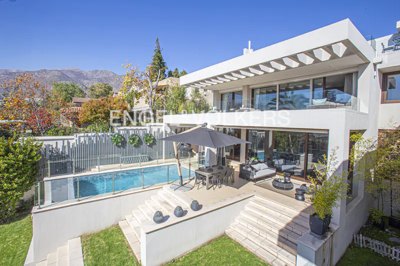 Modern white house with a pool, patio, and mountain view. The patio has an umbrella, table, and chairs. The pool is surrounded by a glass fence.