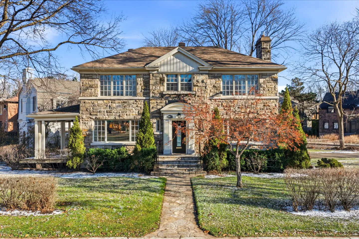 Two-story stone house with a brown roof, surrounded by green lawn and trees under a blue sky. A stone walkway leads to the front door.