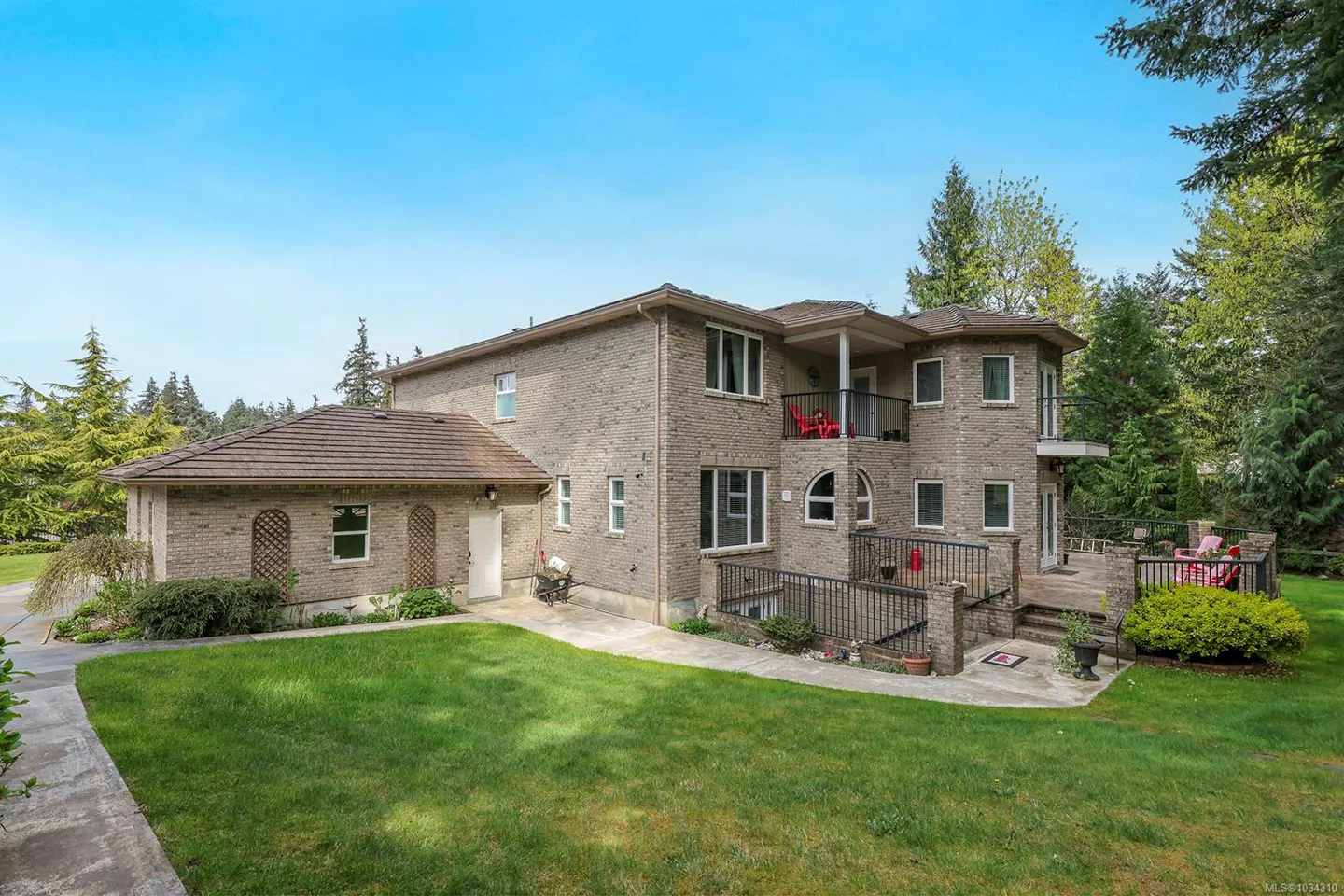 Back view of a two-story brick house with a balcony and a wooden deck, surrounded by green lawn and trees.