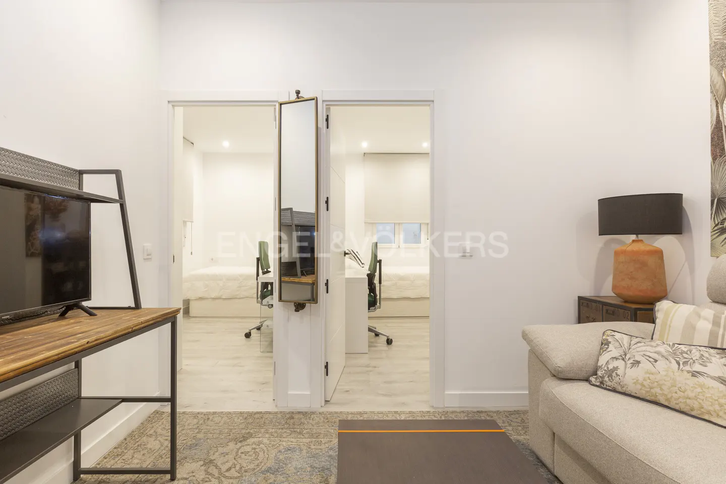 Living room with a TV stand, a sofa, and two open doors leading to bedrooms with desks and beds.