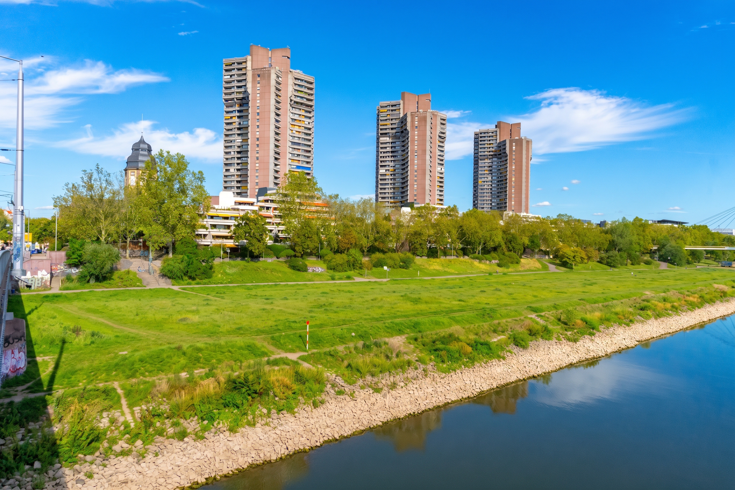 Three tall brown apartment towers rise above a green park and riverbank under a bright blue sky. [AI-generated text]