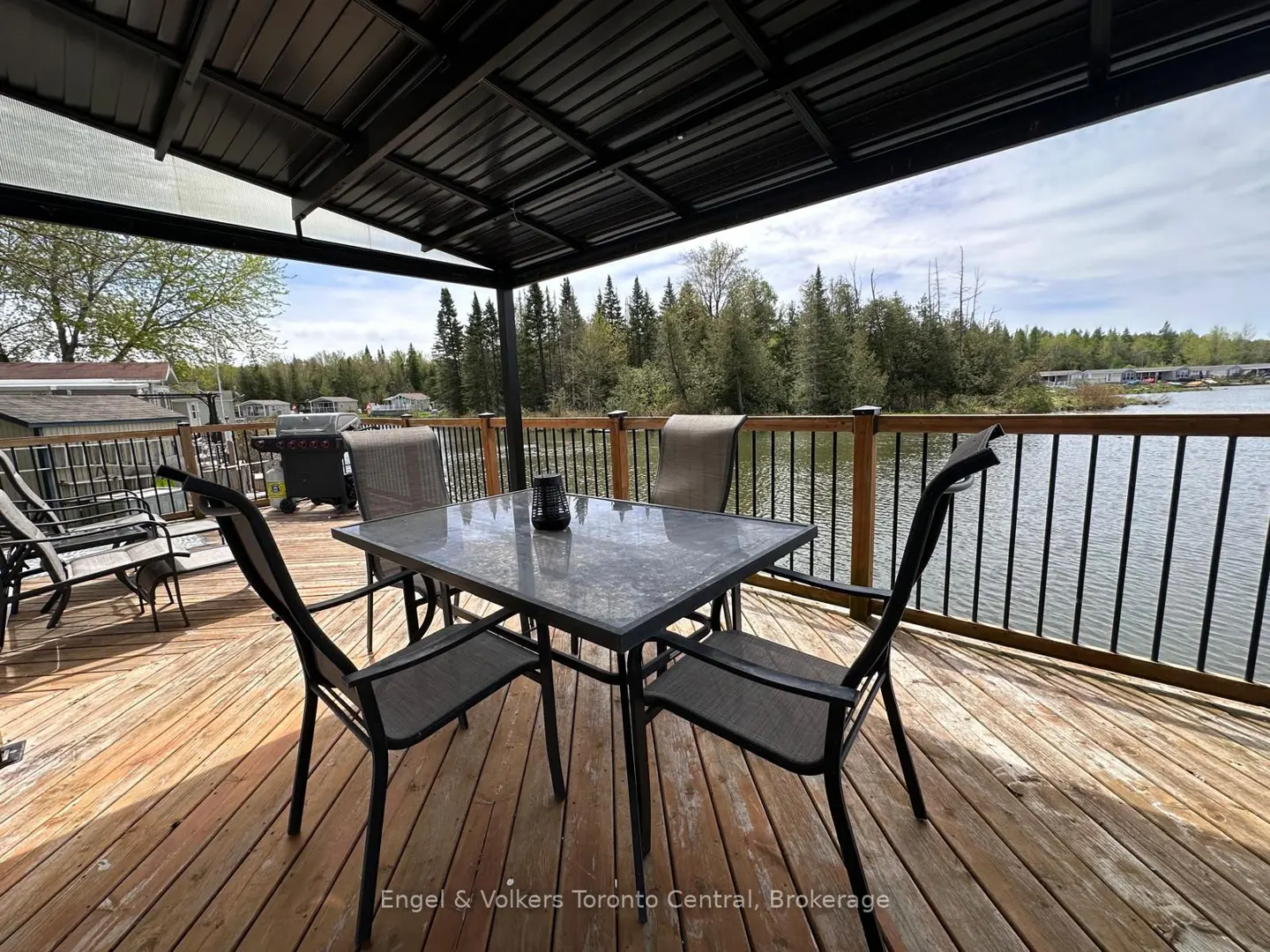 Outdoor patio with a table and chairs on a wooden deck overlooking a lake and trees. The patio has a black metal roof.