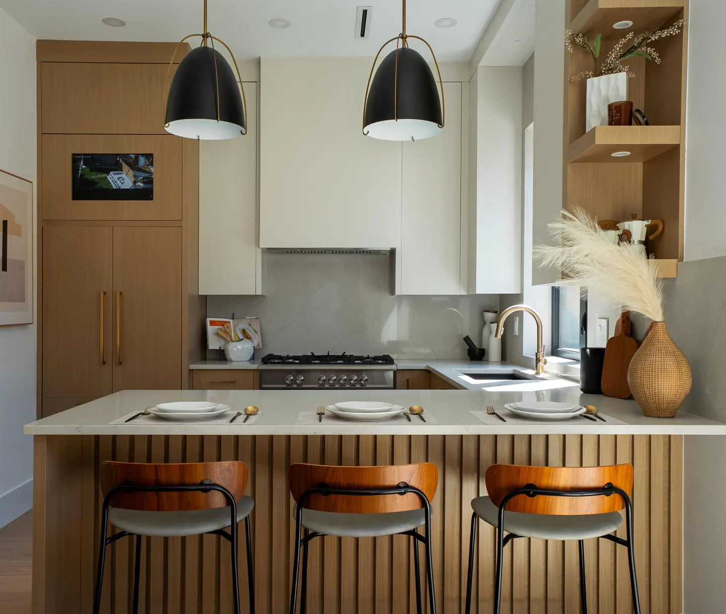A modern kitchen with a breakfast bar, three wooden chairs, and black pendant lights. The cabinets are light wood and white.