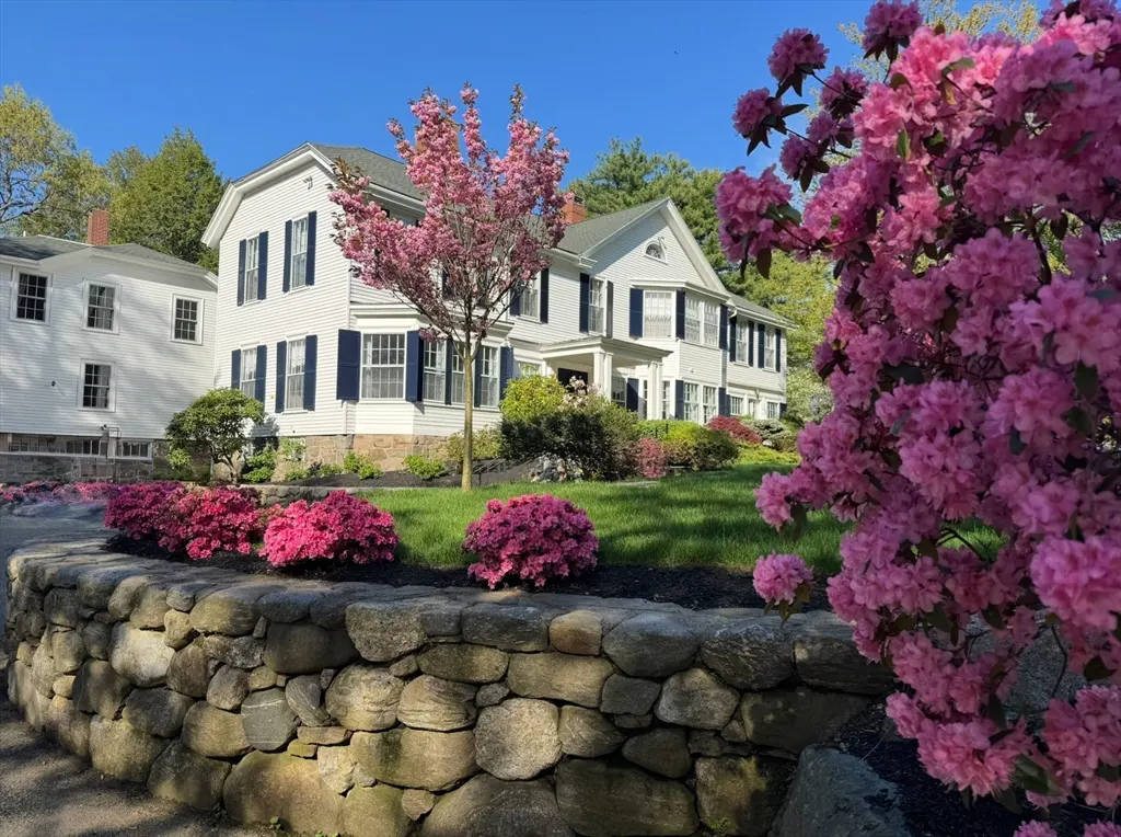 A white two-story house with dark blue shutters, pink flowers, and a stone wall in front.