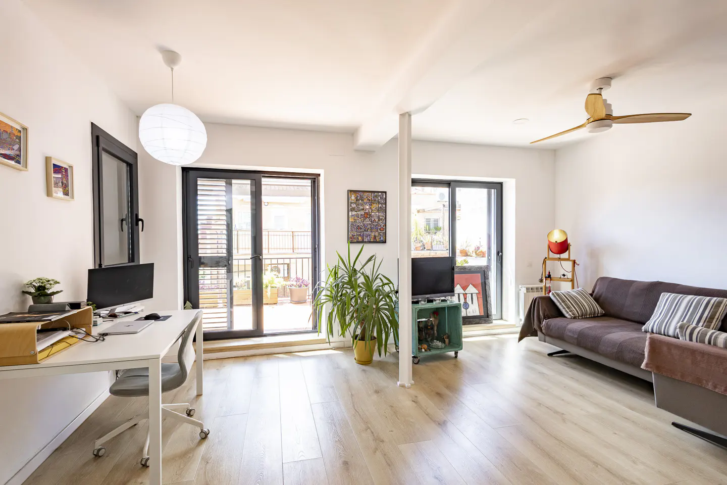 Bright living room with a white desk, brown sofa, and sliding doors to a balcony. A ceiling fan and a paper lantern provide light.