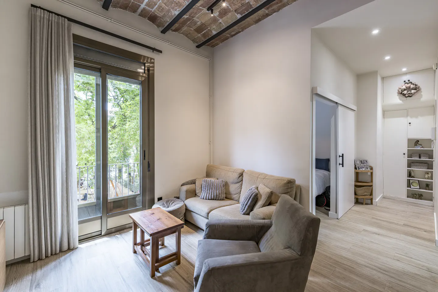 Living room with beige sofa, armchair, wooden coffee table, and sliding door leading to a bedroom under a brick ceiling.