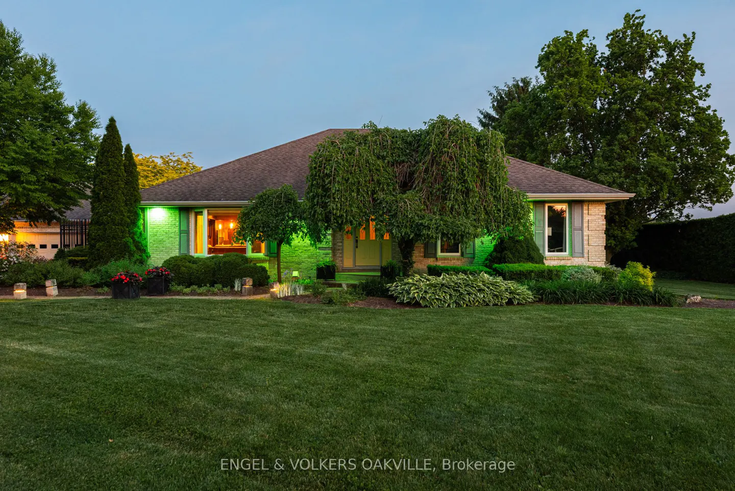 A stone-front bungalow at dusk with a large green lawn, lush landscaping, and warm interior lighting visible through windows.