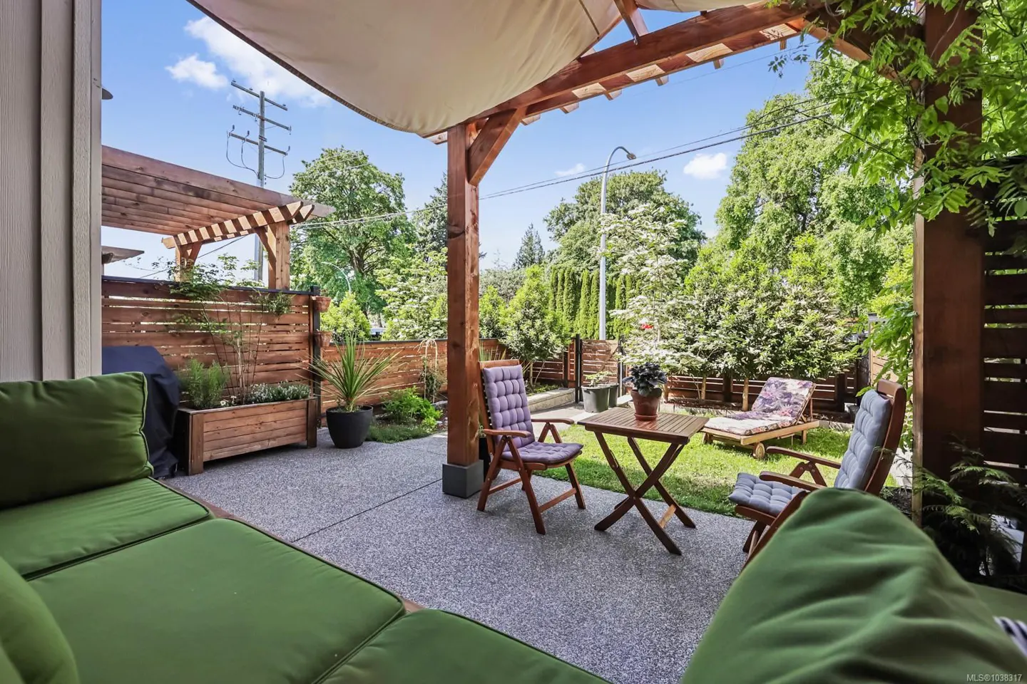Patio with green cushions, wooden pergola, small table, chairs, and a grassy backyard with trees under a blue sky.