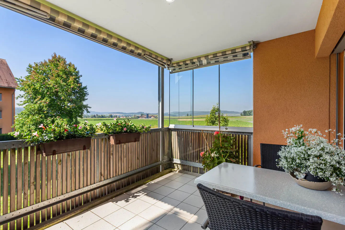 A bright balcony with a table, chairs, and flowers overlooks a green field. The balcony has a wooden railing and glass panels.