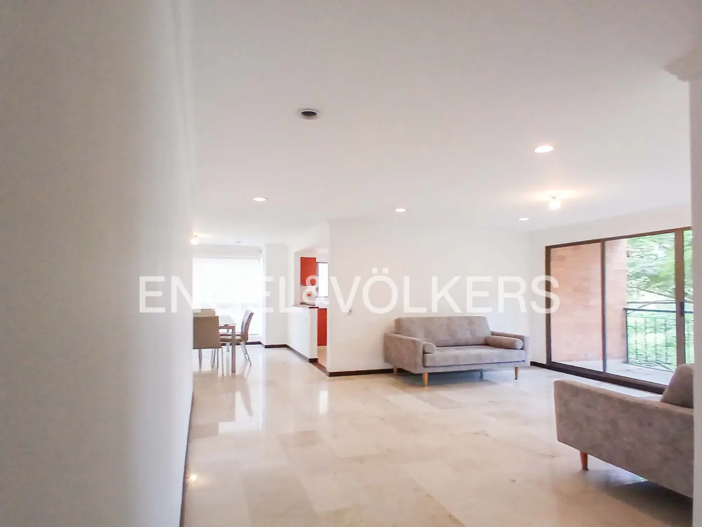 A bright, open-concept living space with marble floors, gray sofas, and a dining area visible in the background. Sliding glass doors lead to a balcony.