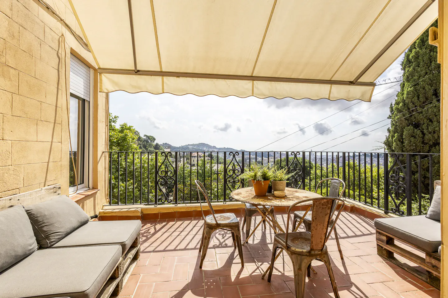 Sunny terrace with a beige awning, pallet sofa, metal table and chairs, overlooking a scenic hillside view.