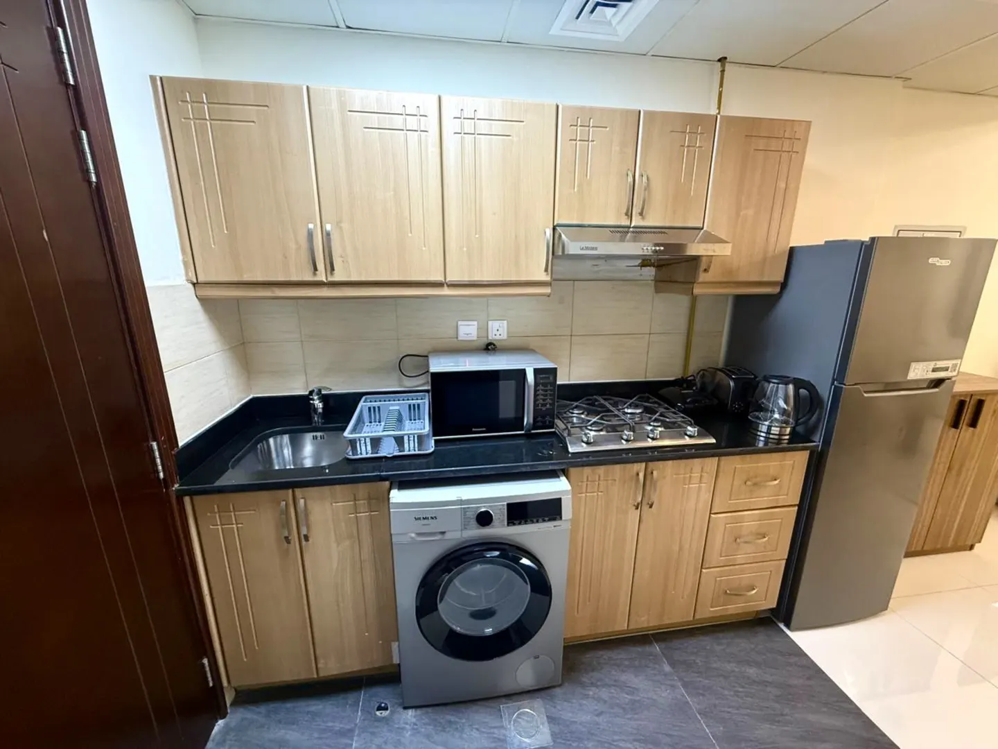A compact kitchen with light wood cabinets, black countertops, and stainless steel appliances including a fridge, microwave, and washing machine.