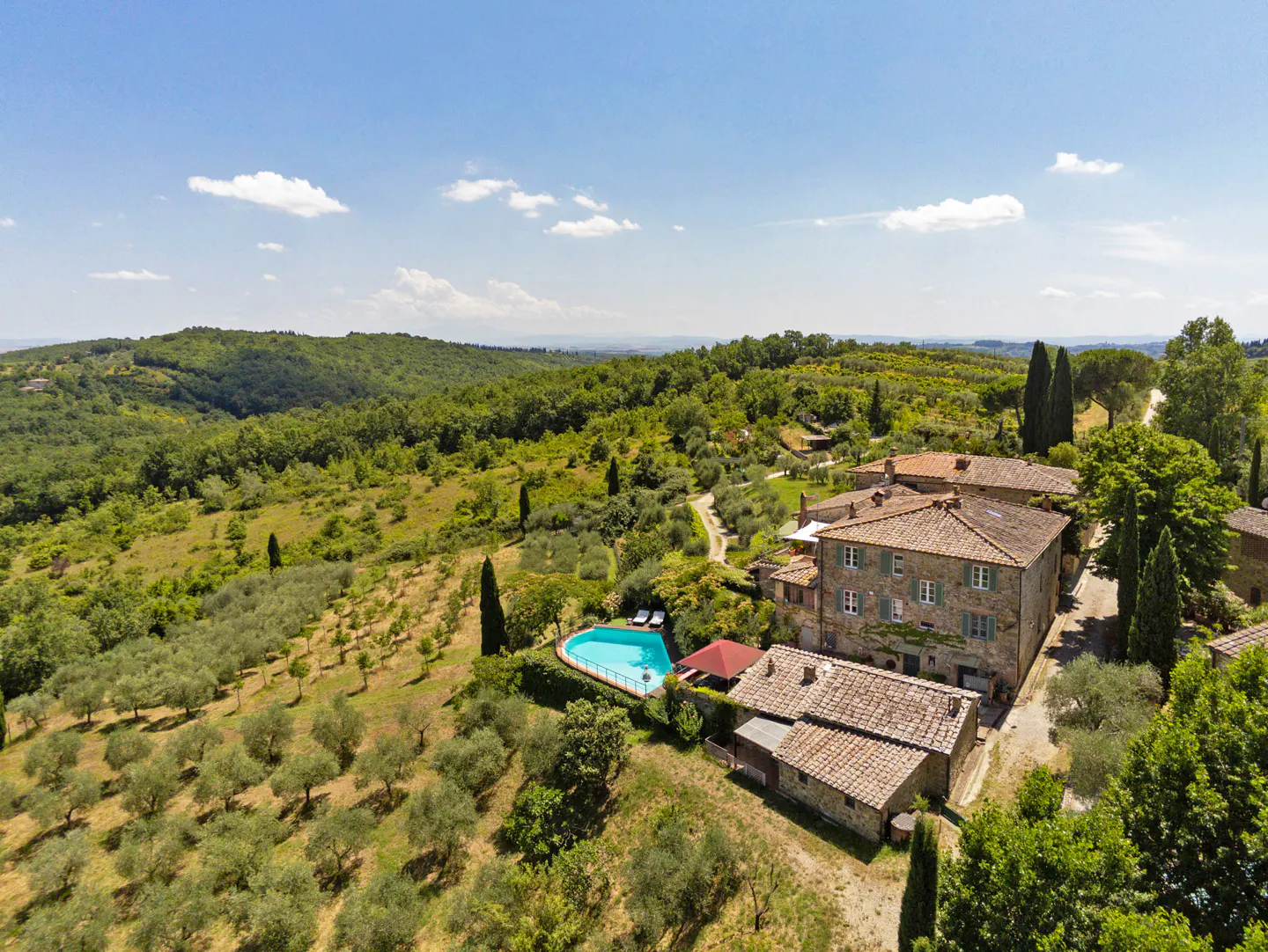 Aerial view of a stone villa with a red tile roof, a blue pool, and olive trees in a green, hilly landscape under a blue sky.