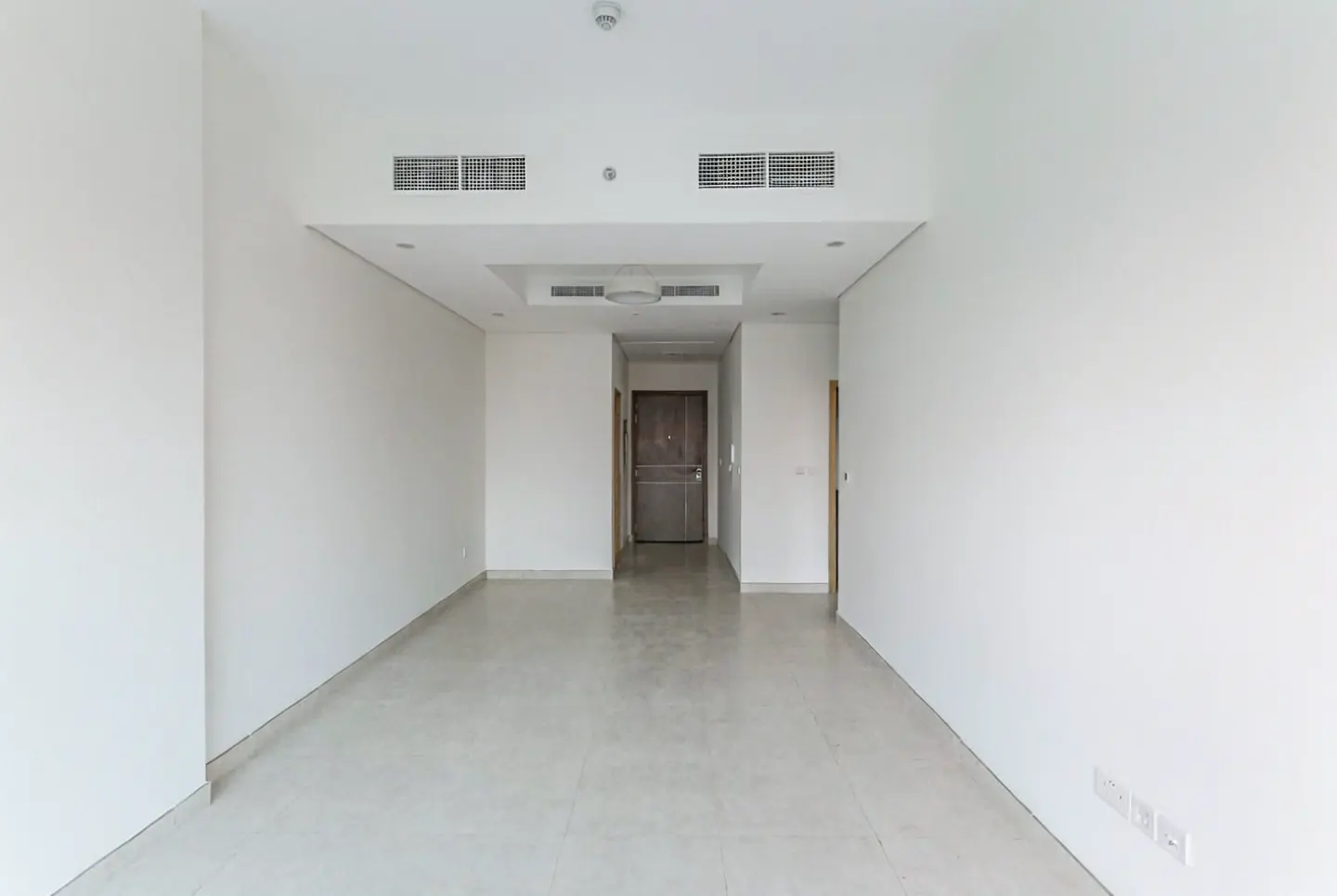 An empty room with white walls and light gray tile flooring. A brown door is visible at the end of the room.