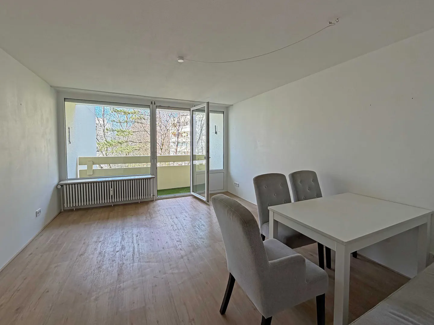 Bright, empty room with wood floors, white walls, and a balcony. A white table and gray chairs sit in the corner.