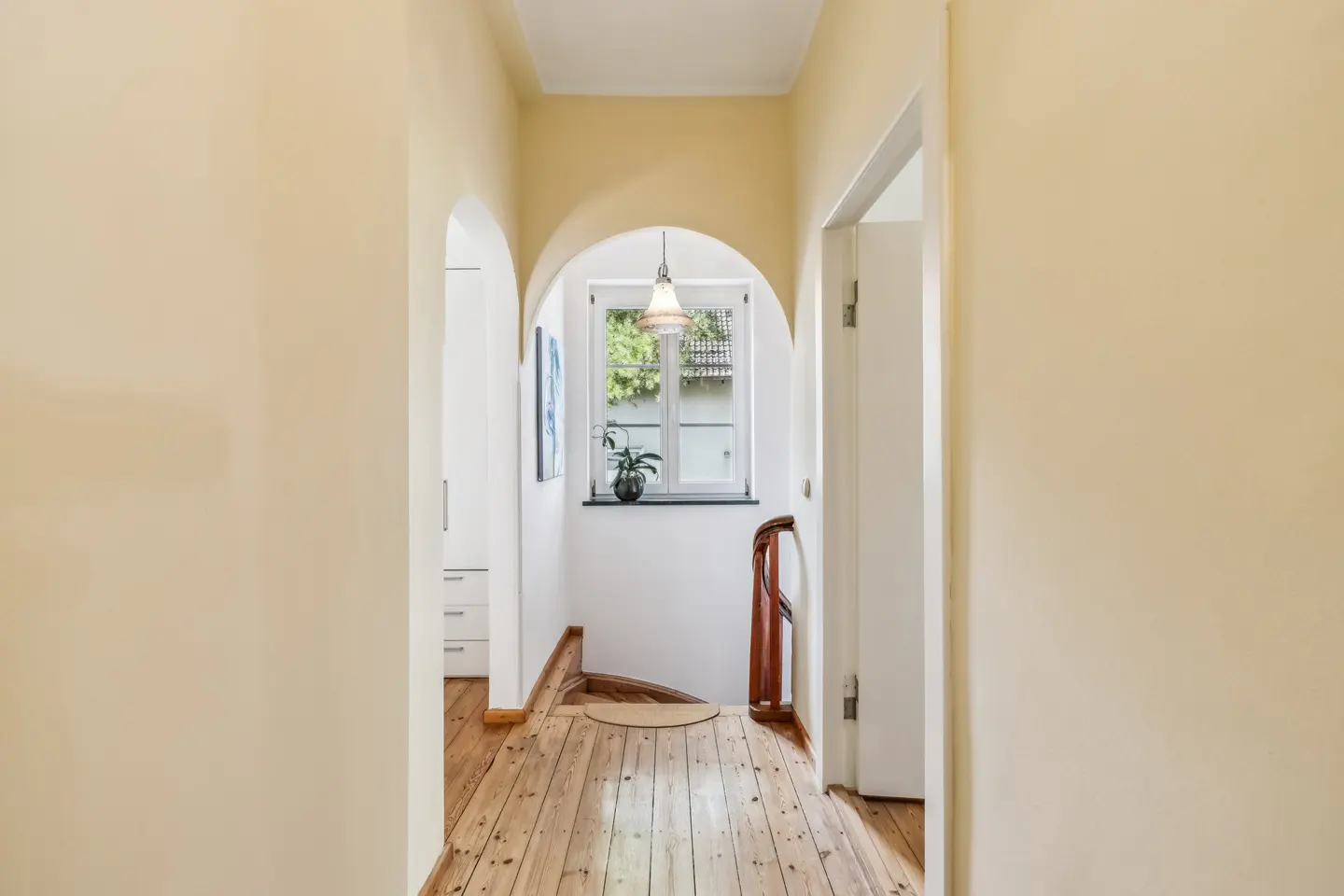 Hallway with arched doorways, light yellow walls, and wood floors. A window with a hanging light is at the end of the hall.