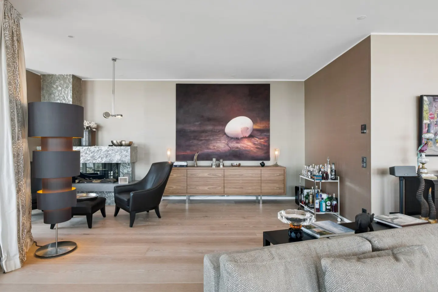 Modern living room with a black armchair, marble fireplace, wooden sideboard, large egg art, and a bar cart on wood floors.