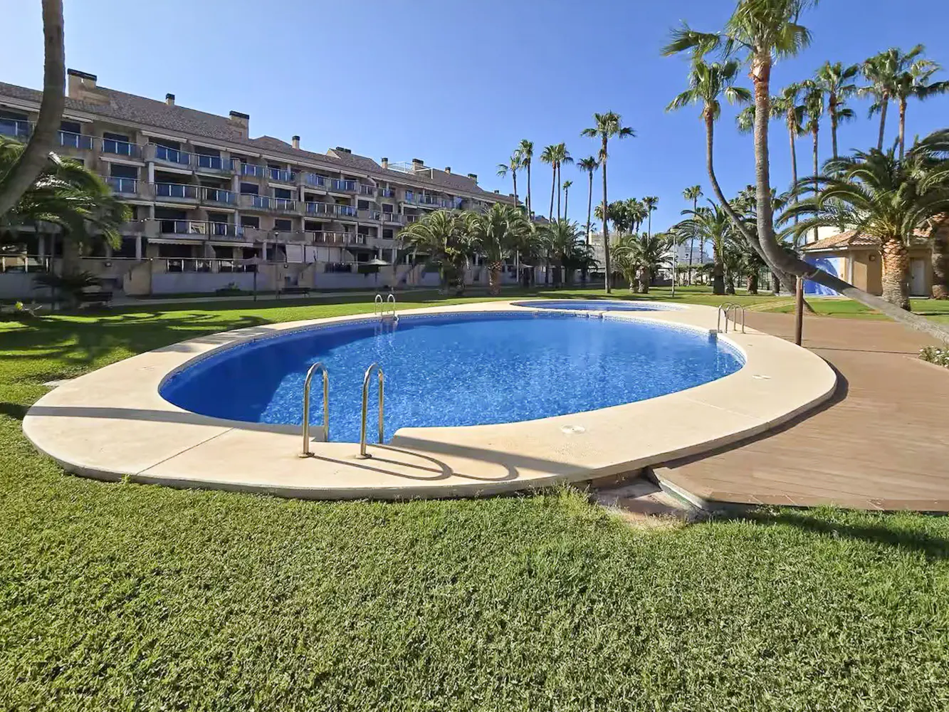 Outdoor pool with blue water and metal ladder, surrounded by green grass, palm trees, and a multi-story building under a clear blue sky.
