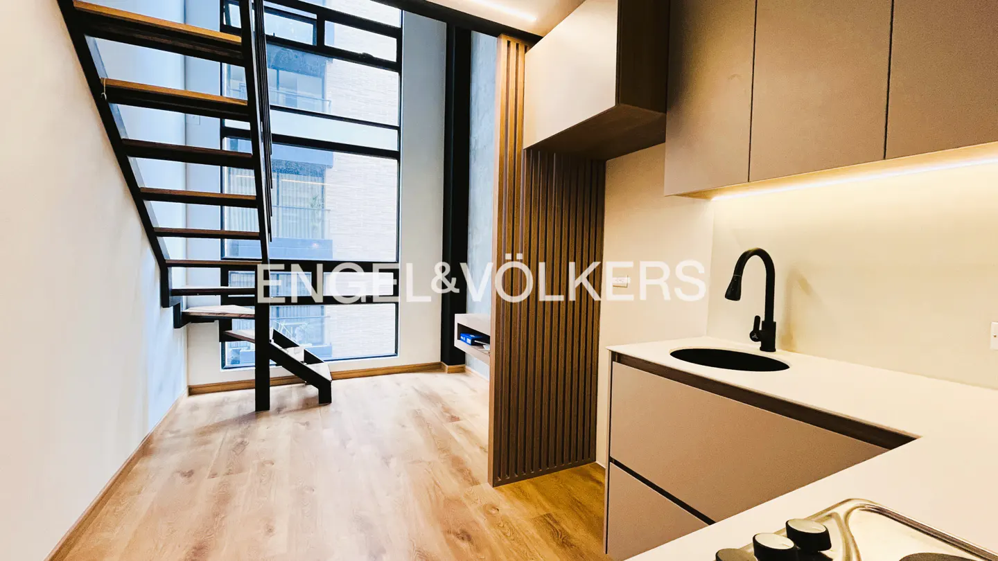 Modern kitchen with light wood floors, sleek cabinets, a black faucet, and a floating staircase by a tall window.