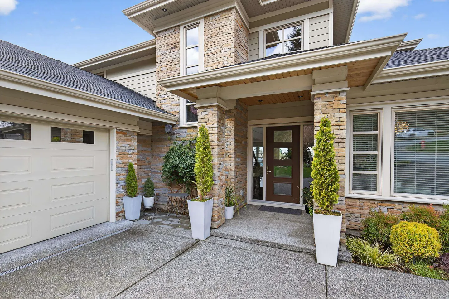 Modern home exterior with stone facade, beige garage door, brown front door, and potted evergreen trees on a concrete path.