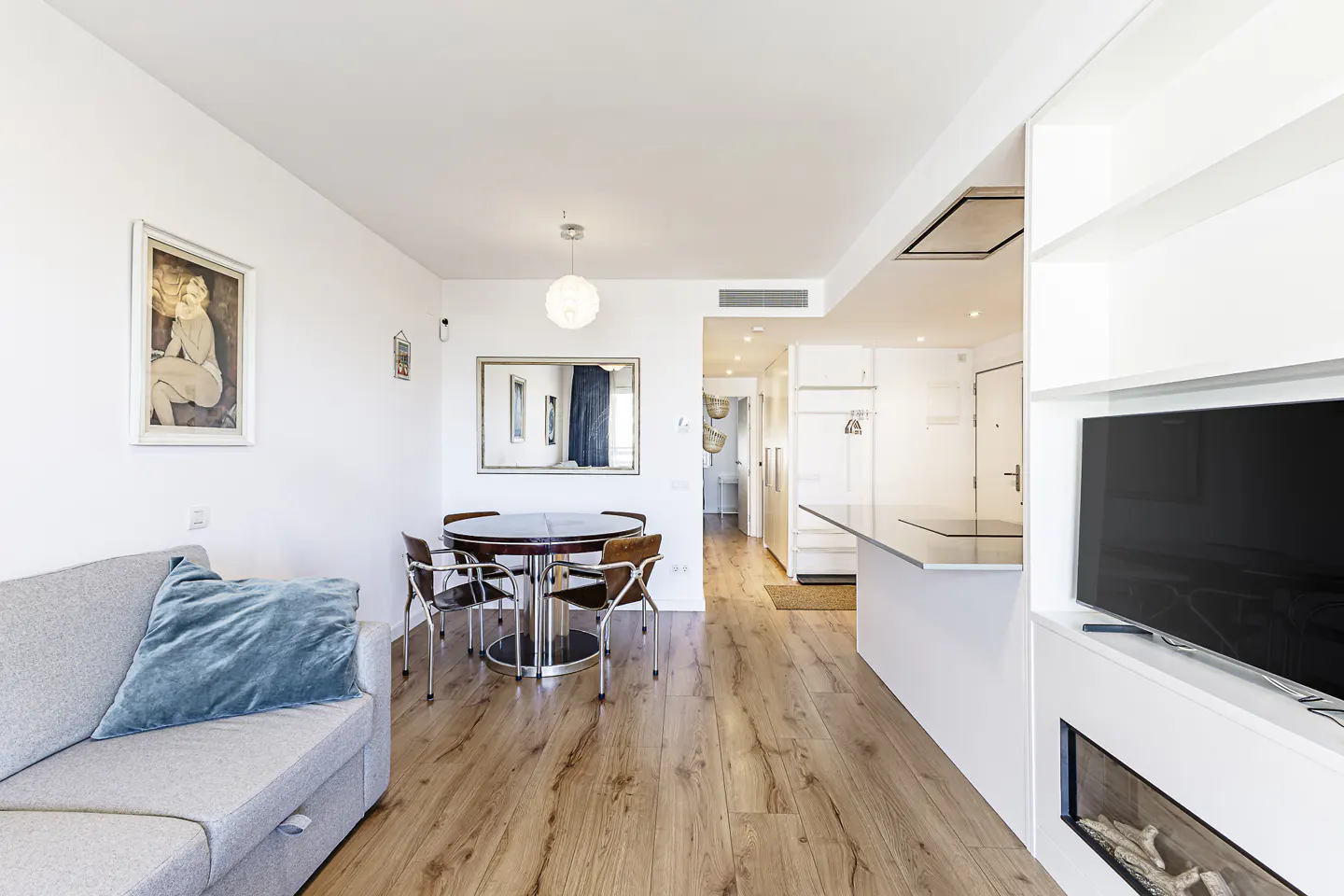 Bright, modern apartment interior with wood floors, a gray sofa with a blue pillow, and a round dining table with four chairs. A TV and fireplace are built into a white wall unit.