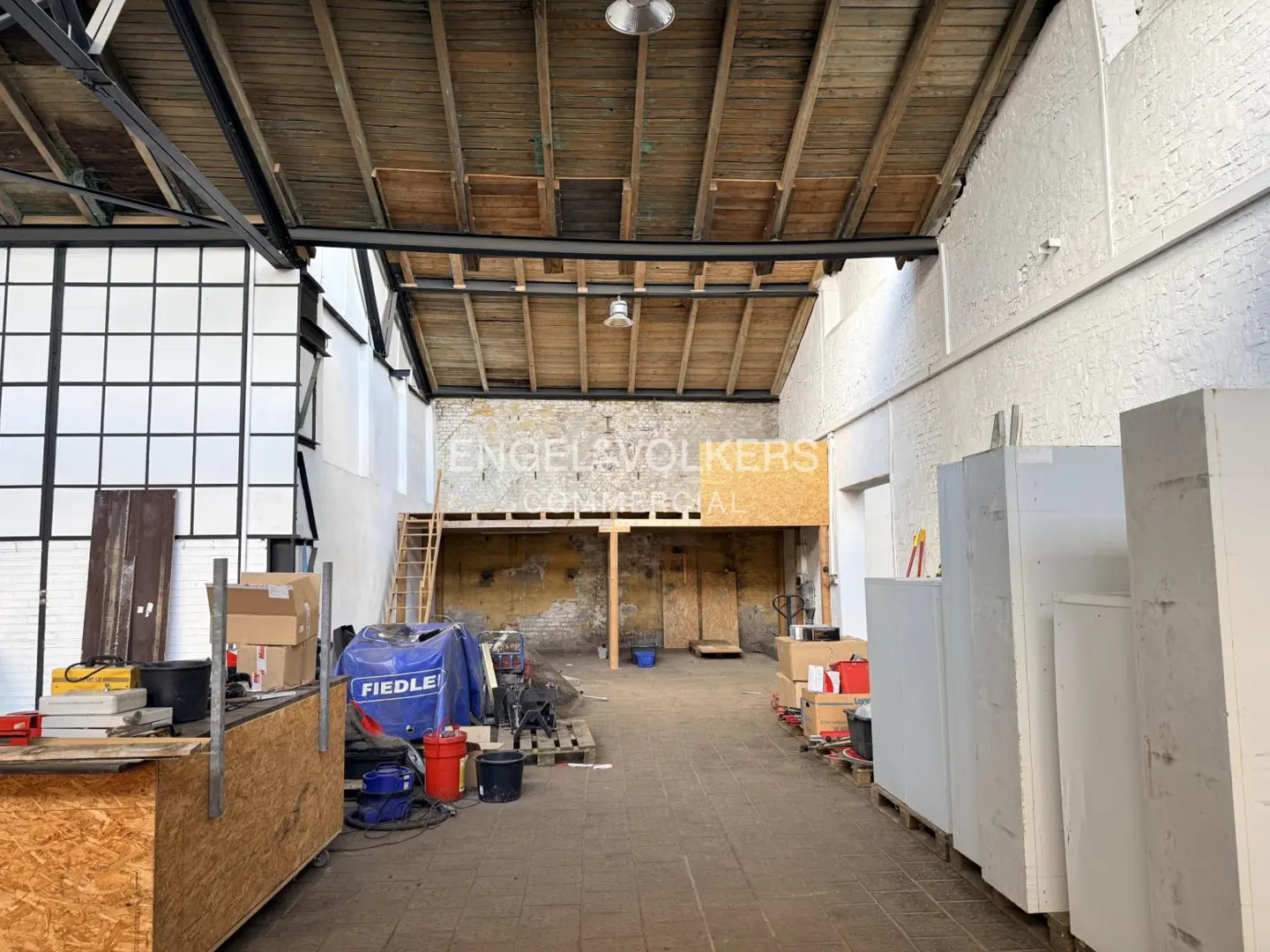 Interior view of a commercial space with a wooden ceiling, white brick walls, and an Engel & Volkers sign. Boxes and equipment are scattered around.