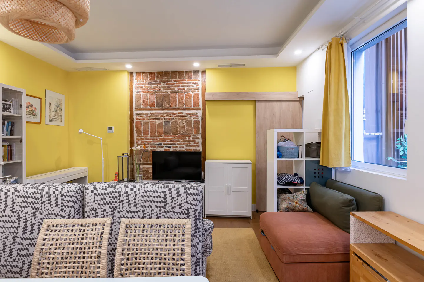 Bright living room with yellow walls, brick accent, and patterned sofa. A woven light fixture hangs from the ceiling.