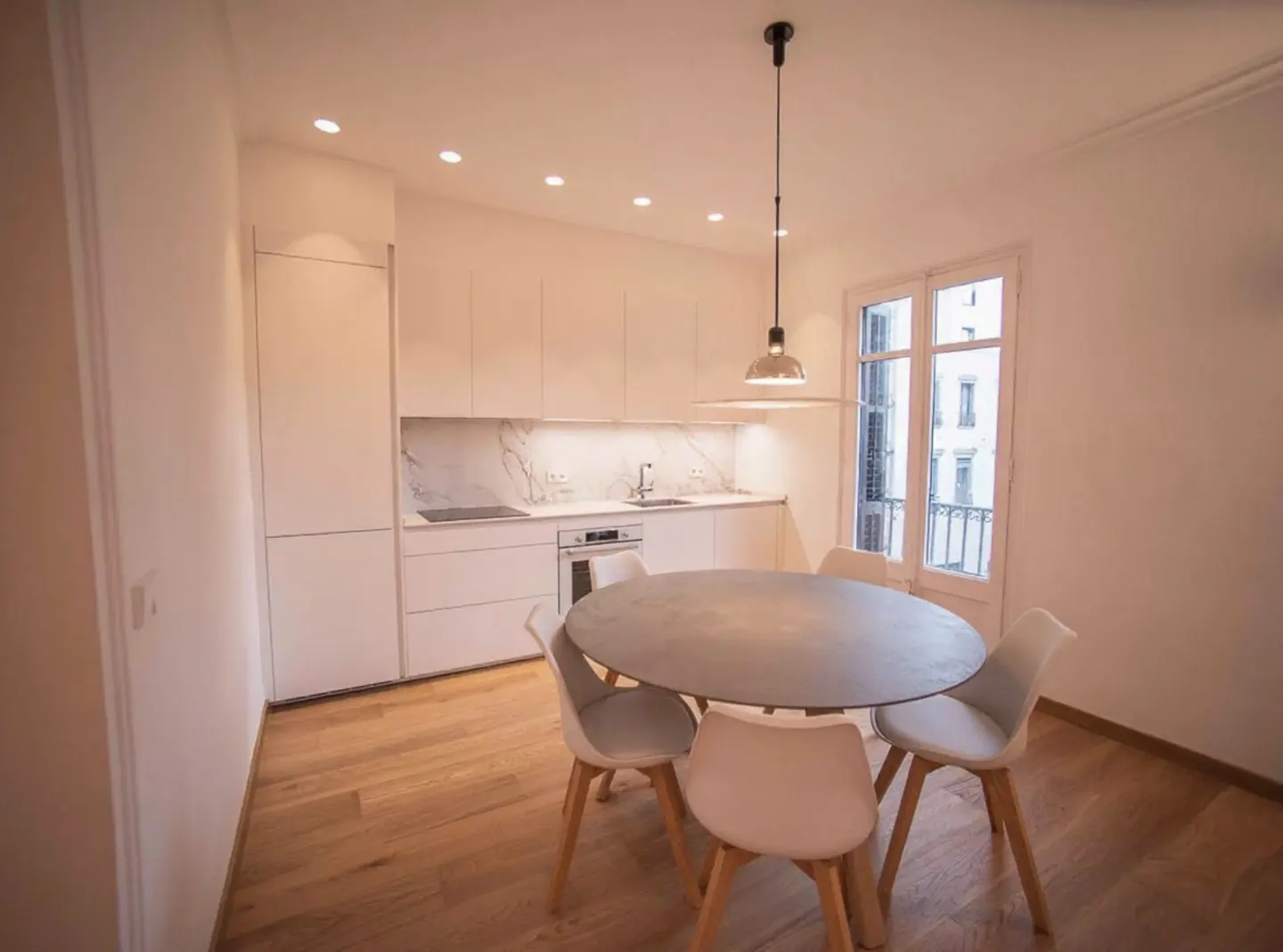 Bright, modern kitchen with white cabinets, marble backsplash, and wood floors. A round table with chairs sits in front of a window.