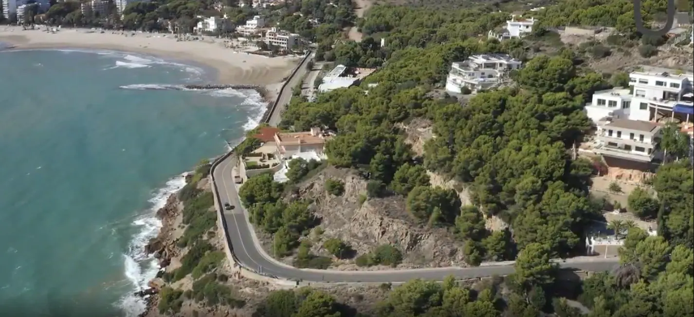 Aerial view of a coastline with a beach, turquoise water, a winding road, and houses on a green, tree-covered hillside.