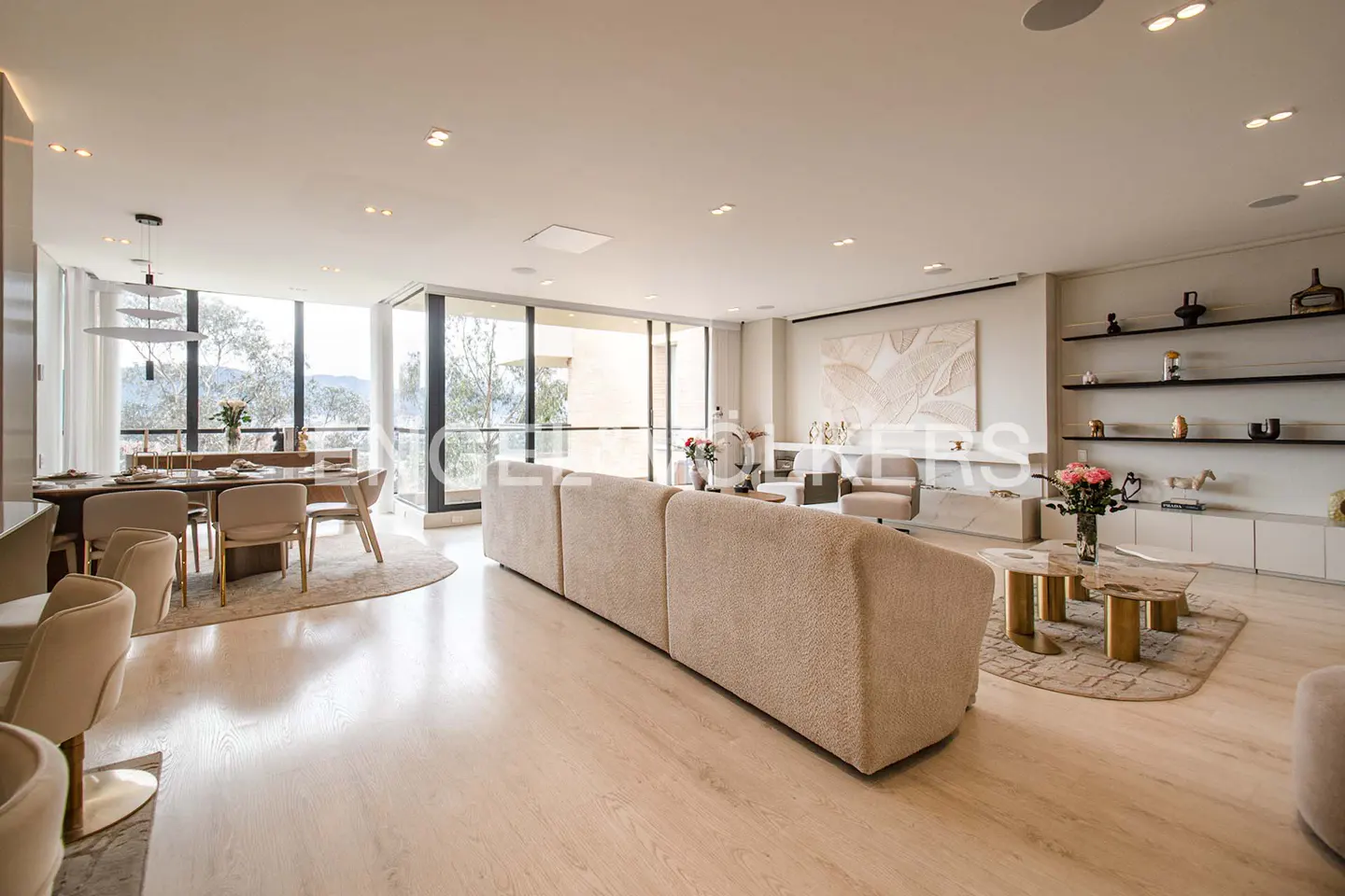 A bright, open-concept living and dining area with beige sofa, wood floors, and large windows overlooking greenery.