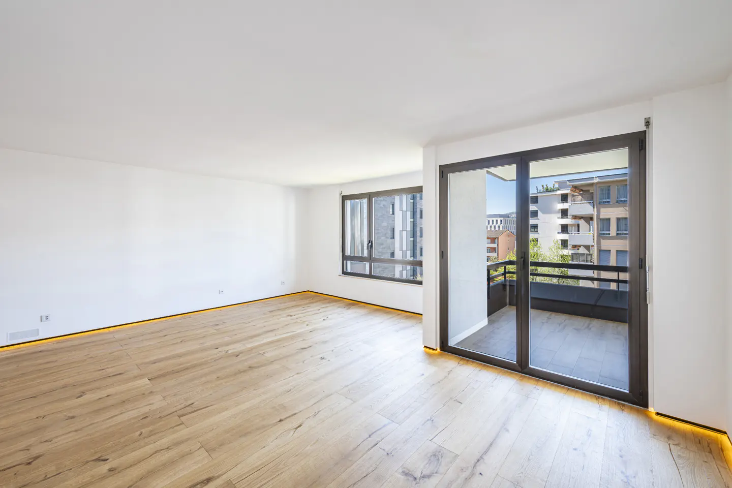 Bright, empty room with light wood floors, white walls, and black-framed windows and sliding glass doors to a balcony.