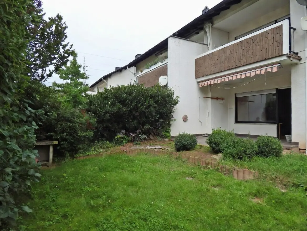 Exterior view of a white apartment building with a green lawn and bushes in front. Balconies with wood railings are visible.