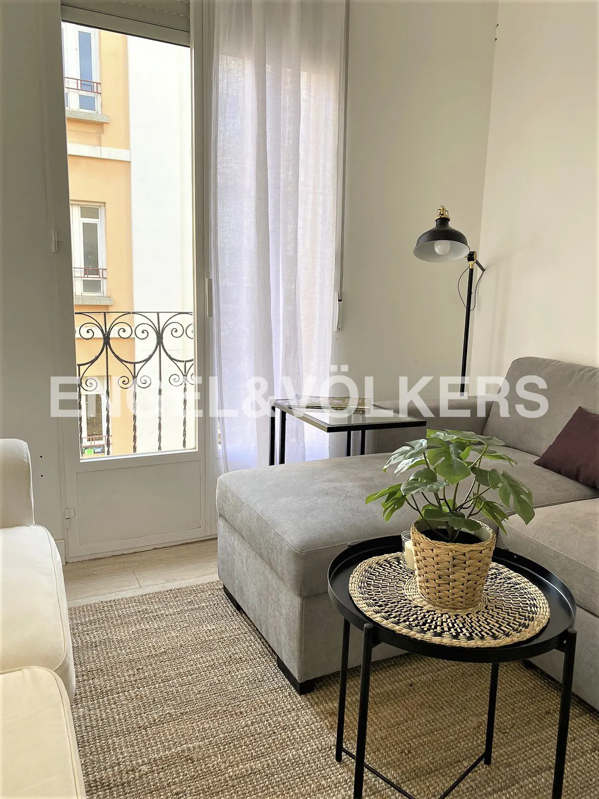 Living room with a gray sofa, black lamp, and a round table with a plant. Balcony doors with sheer white curtains are open.