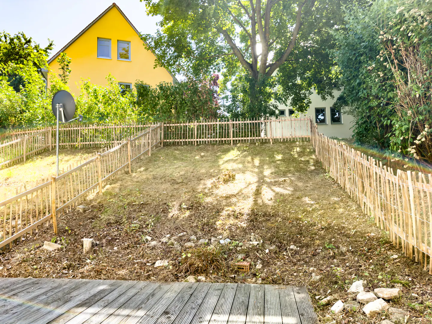 A wooden deck overlooks a sloped, fenced backyard with a bright yellow house in the background under a sunny sky.