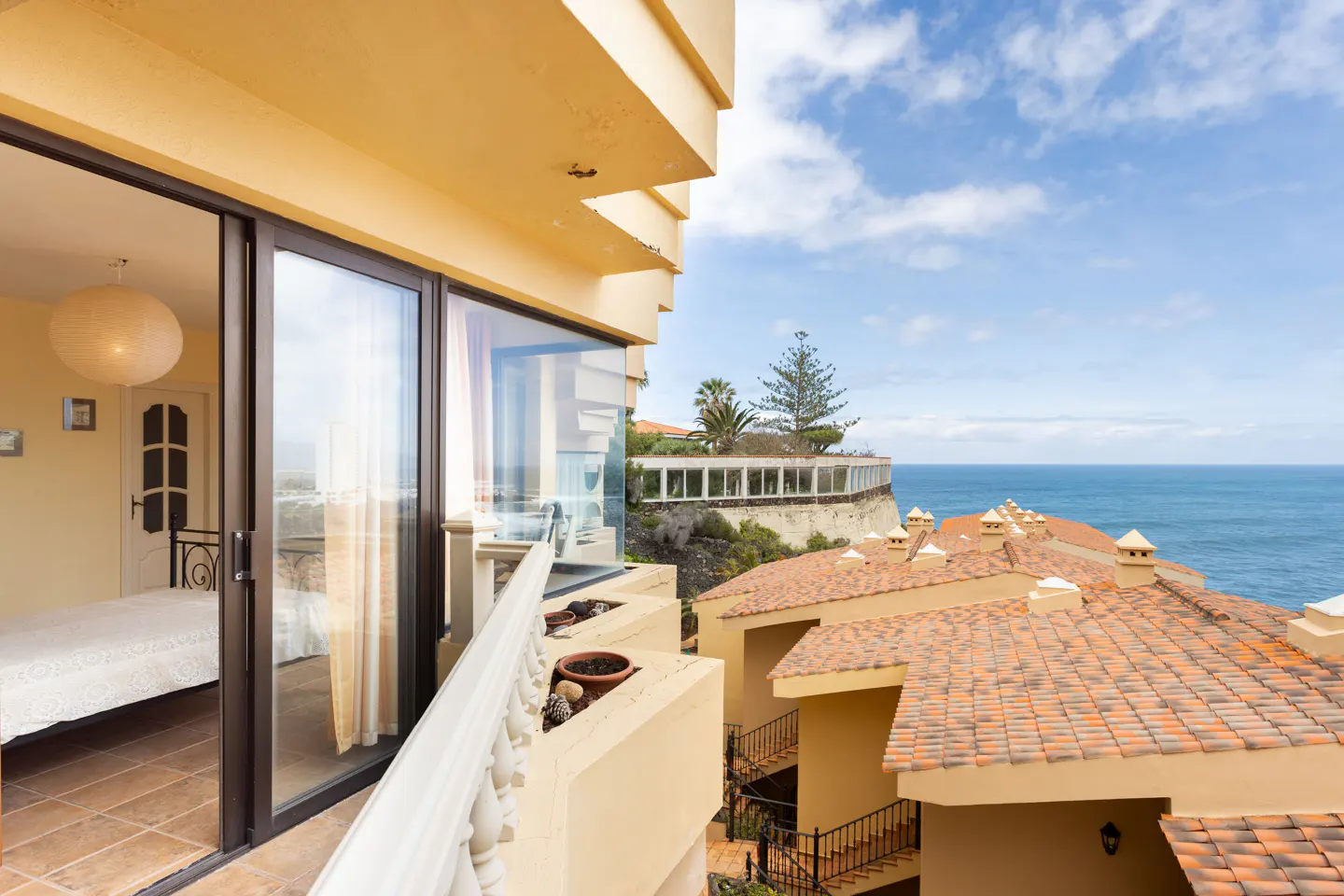View from a balcony with a bedroom visible through sliding glass doors, overlooking terracotta rooftops and the ocean.