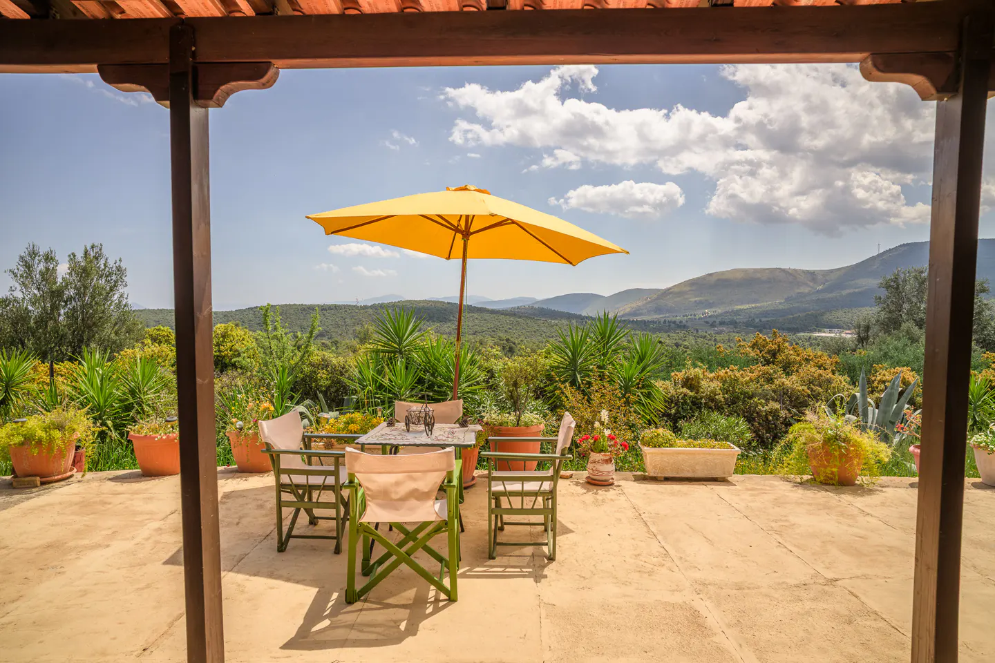 Outdoor patio with table, chairs, and yellow umbrella overlooking green hills under a blue sky with clouds.