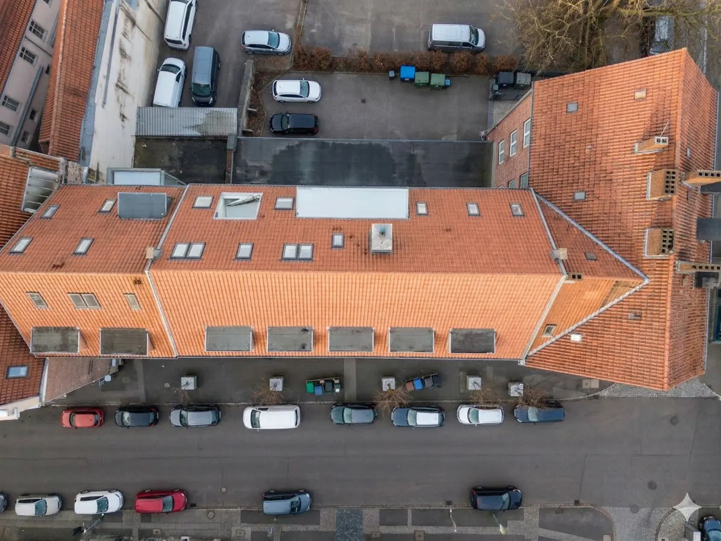 Aerial view of a building with a red tile roof, skylights, and parked cars on the street and in the parking lot.