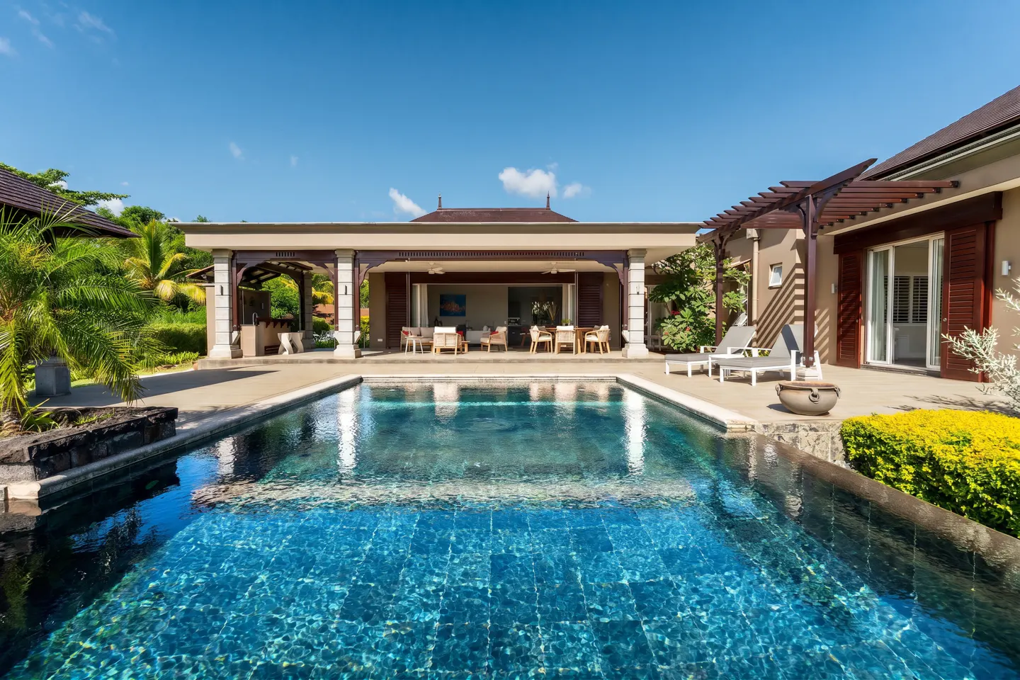 A luxury home's pool reflects the blue sky. Patio furniture sits under a covered porch. Lounge chairs are on the deck.