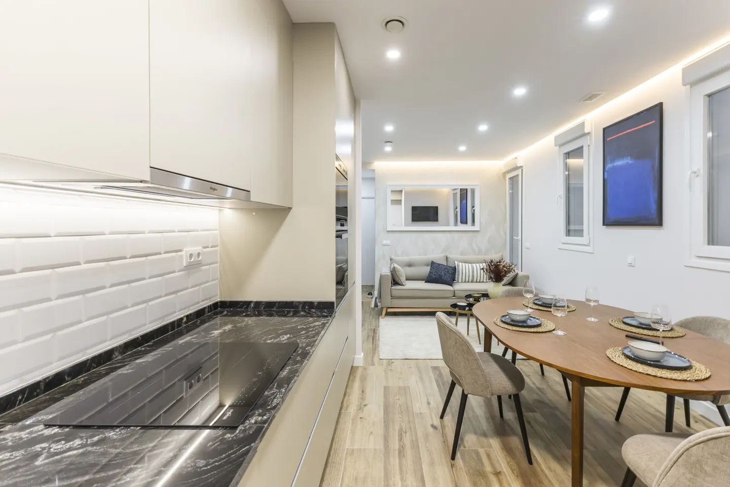 Bright, modern apartment interior with kitchen, dining table, sofa, and artwork. White walls and wood floors create a clean, open space.