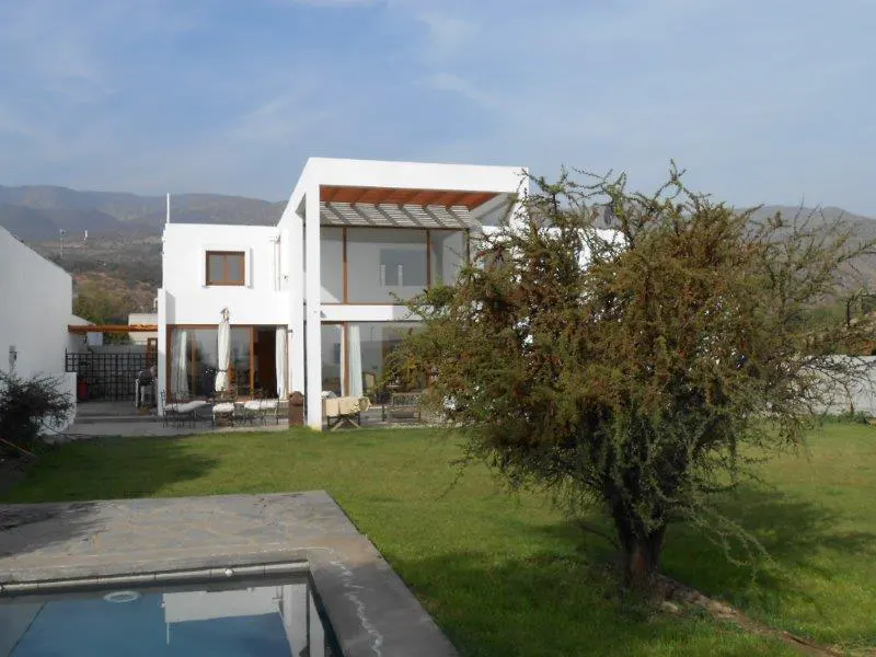 Modern white house with large windows, a pool, and a tree in the yard. Mountains are visible in the background.