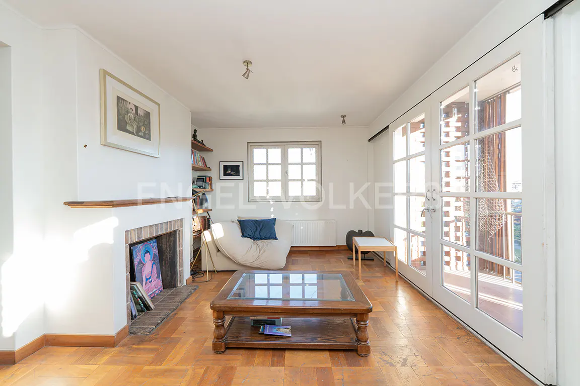Bright living room with hardwood floors, white walls, fireplace, and glass doors to a balcony. A sofa, coffee table, and bookshelf are also visible.