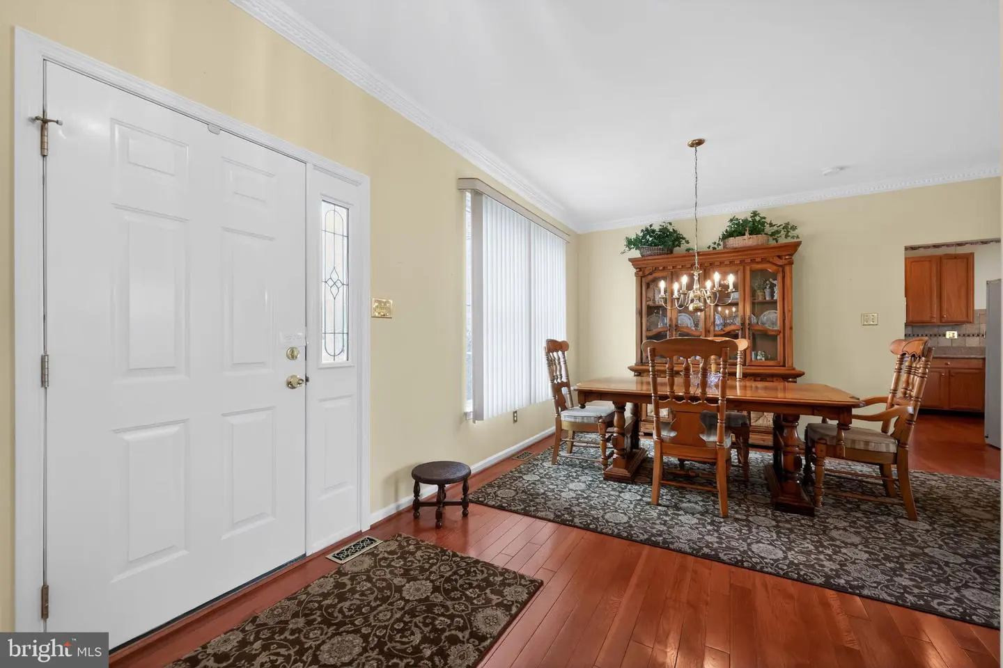 Entryway with white door, wood floors, and dining area. Table, chairs, and hutch on a patterned rug.