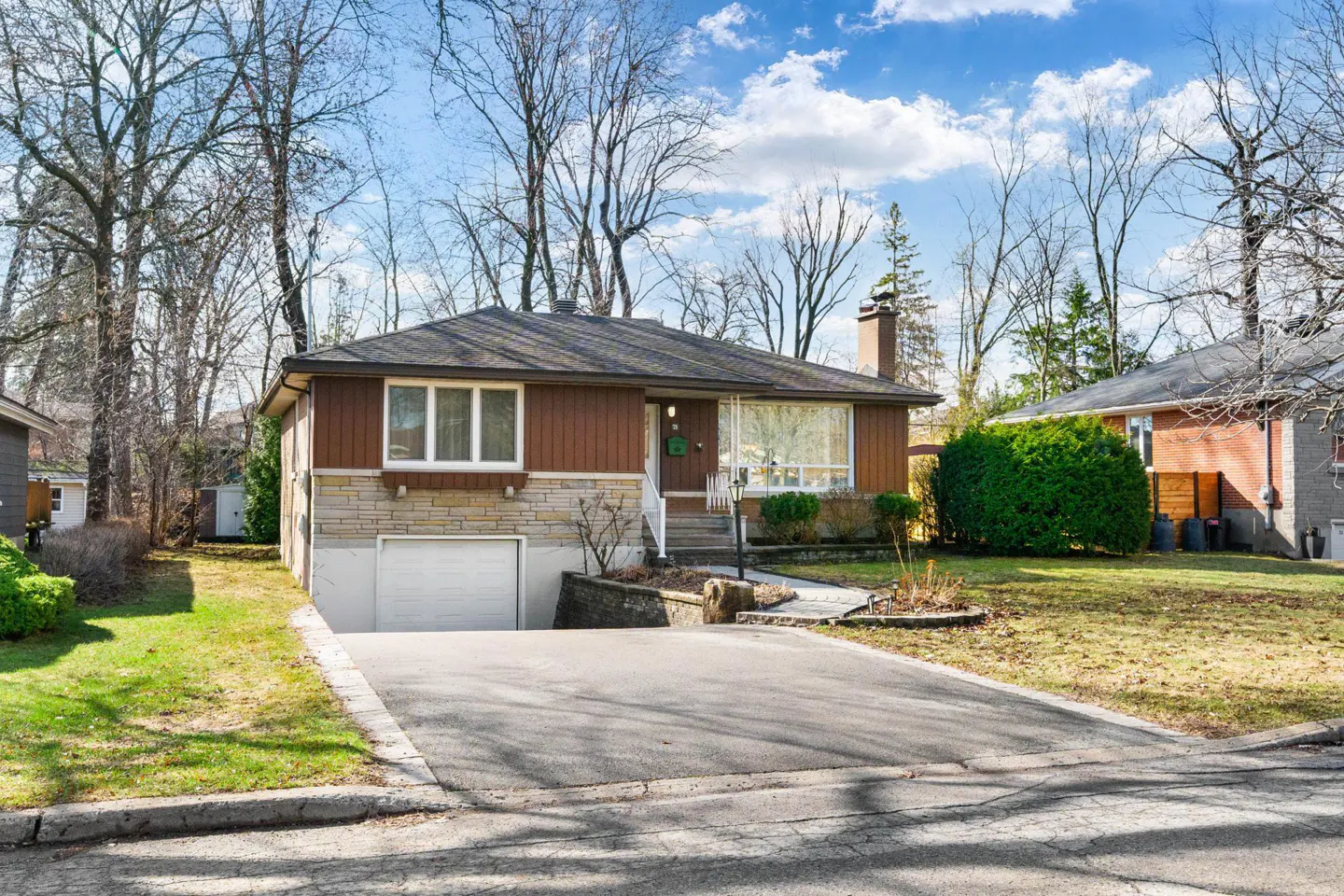A single-story house with a brown facade, stone accents, and a driveway leading to a garage. Bare trees surround the property.