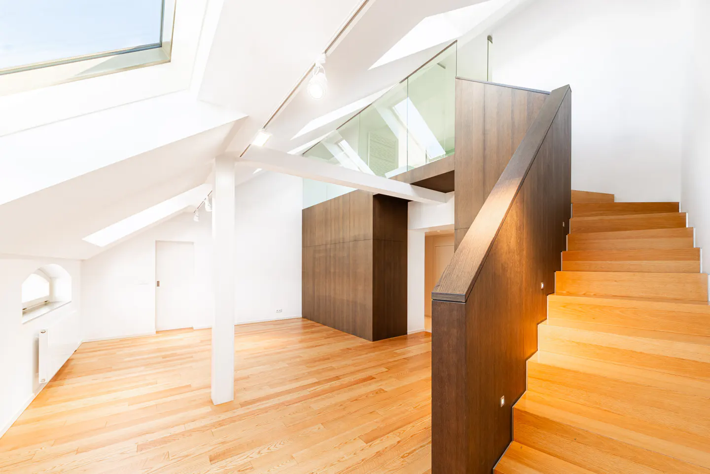 Bright, modern interior with light wood floors, white walls, and a wooden staircase with dark brown trim. Skylights provide natural light.