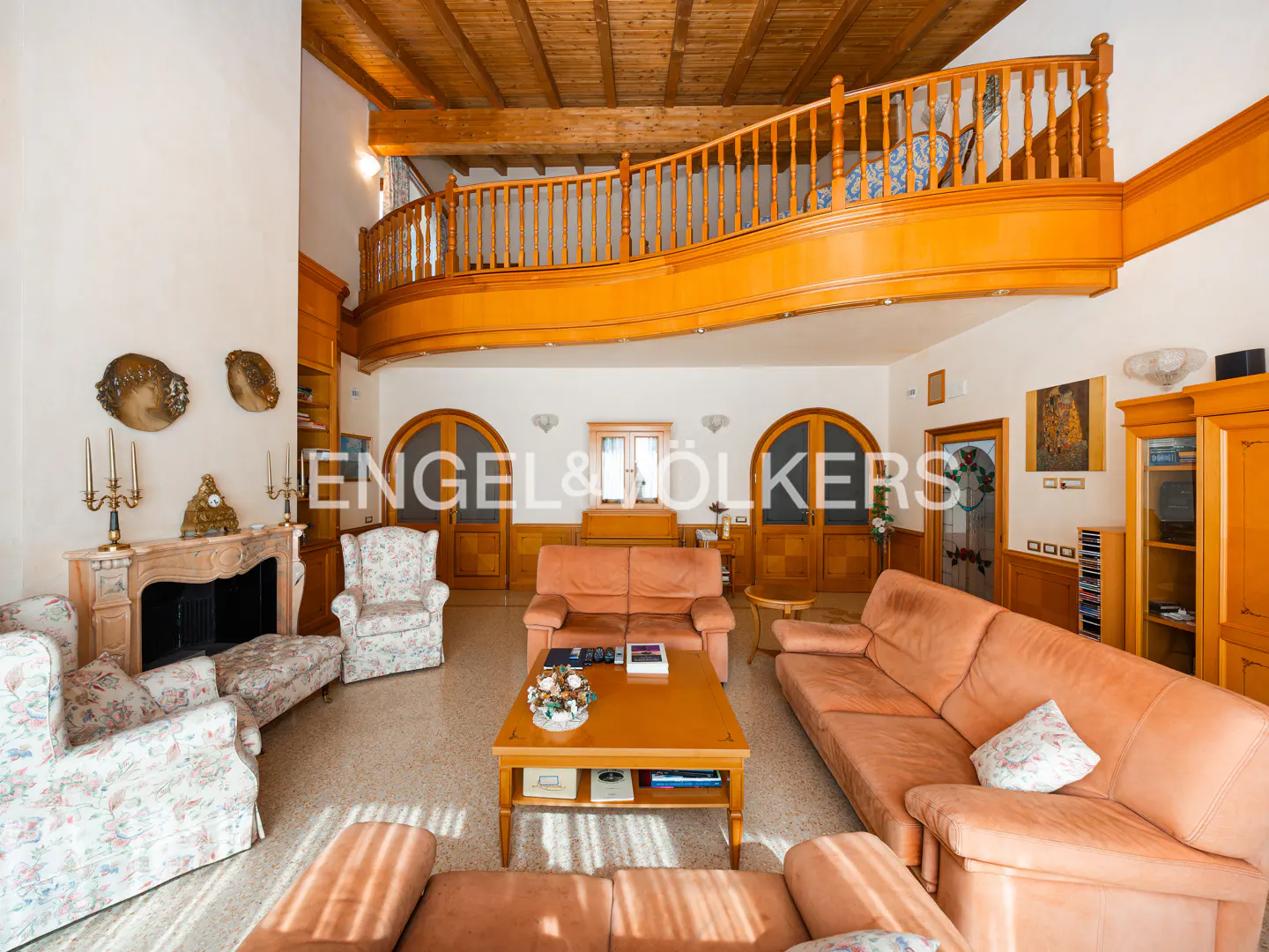 A spacious living room with a wooden balcony, sofas, a fireplace, and arched doors.