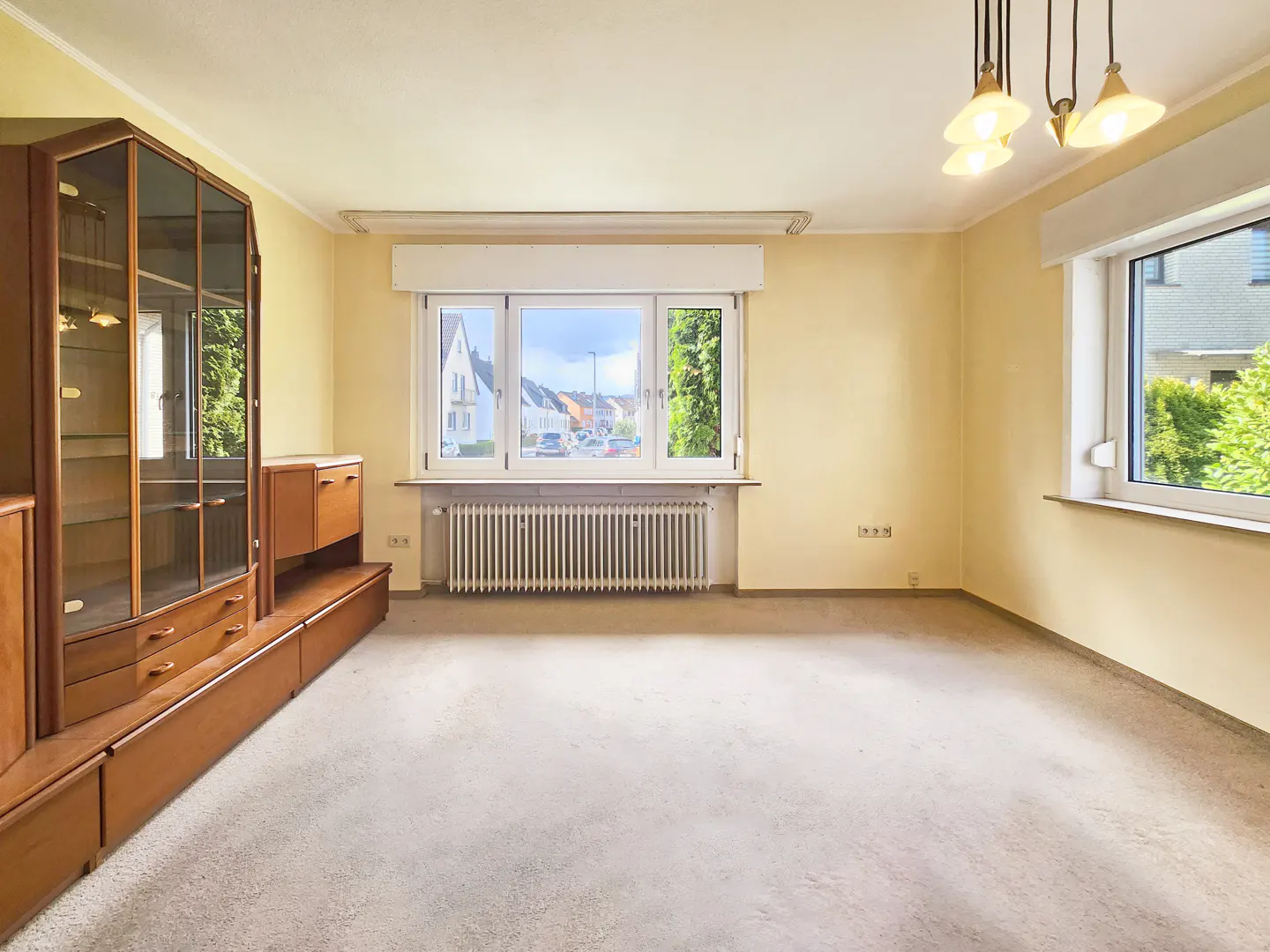 Empty room with beige carpet, yellow walls, and a large wooden cabinet. Windows show a residential street and greenery. A radiator sits under the main window.