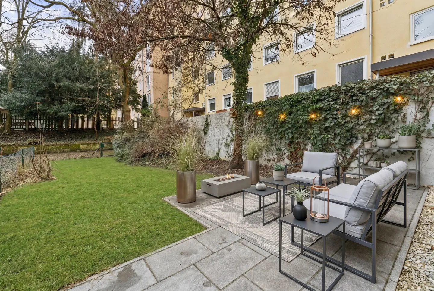 A backyard patio with gray furniture, a fire pit, and a green lawn. The patio is surrounded by a wall covered in ivy and string lights.