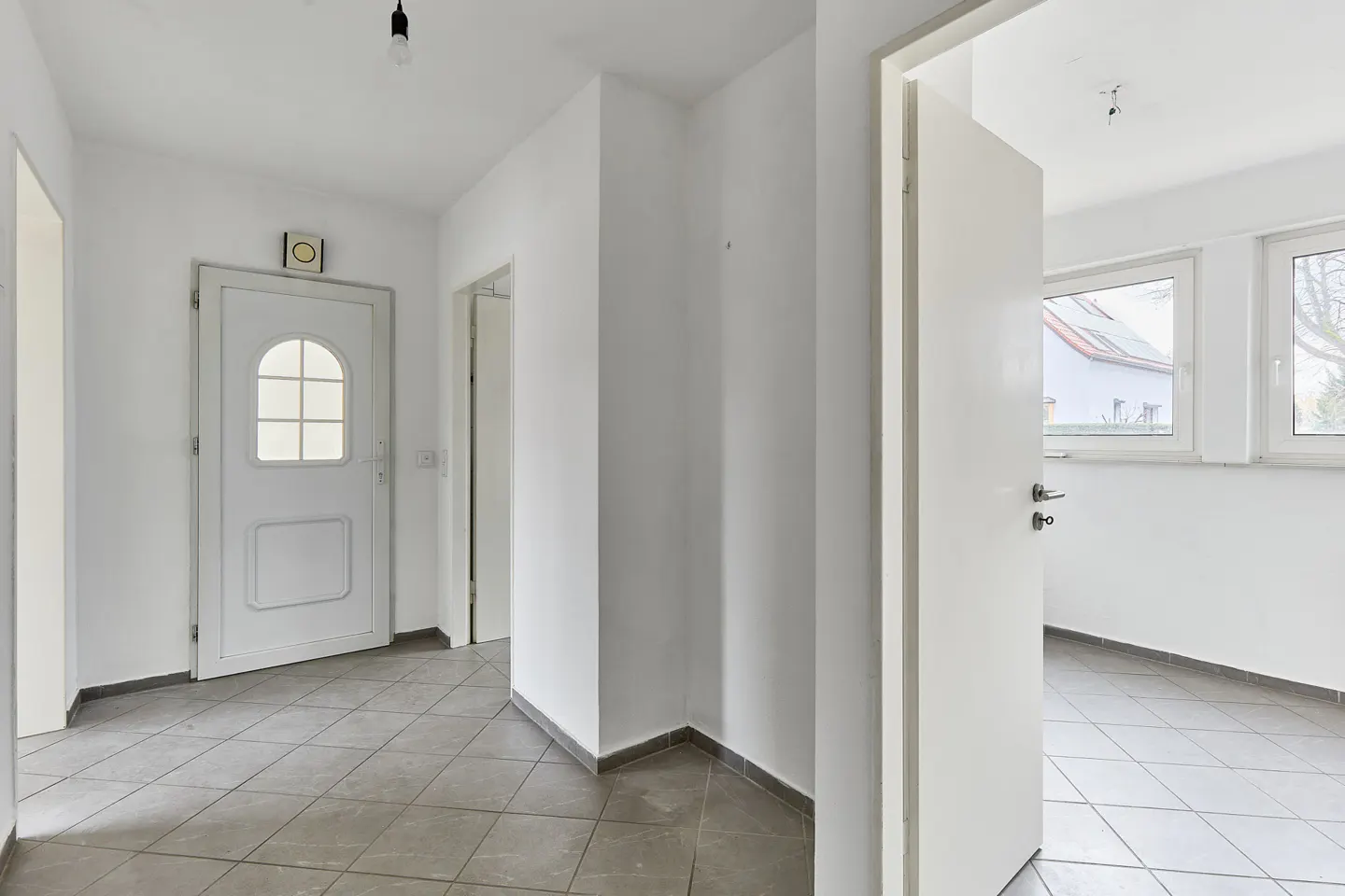 Bright, white hallway with gray tile floor. A white front door with glass panes is on the left. An open doorway leads to a room with windows.