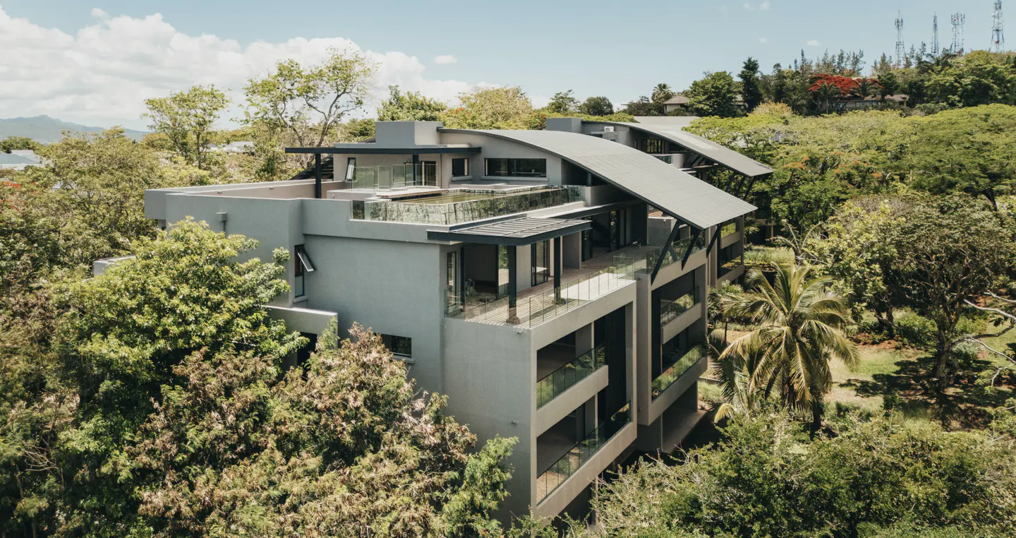 Aerial view of a modern, gray apartment building with curved roofs, surrounded by lush green trees and foliage.
