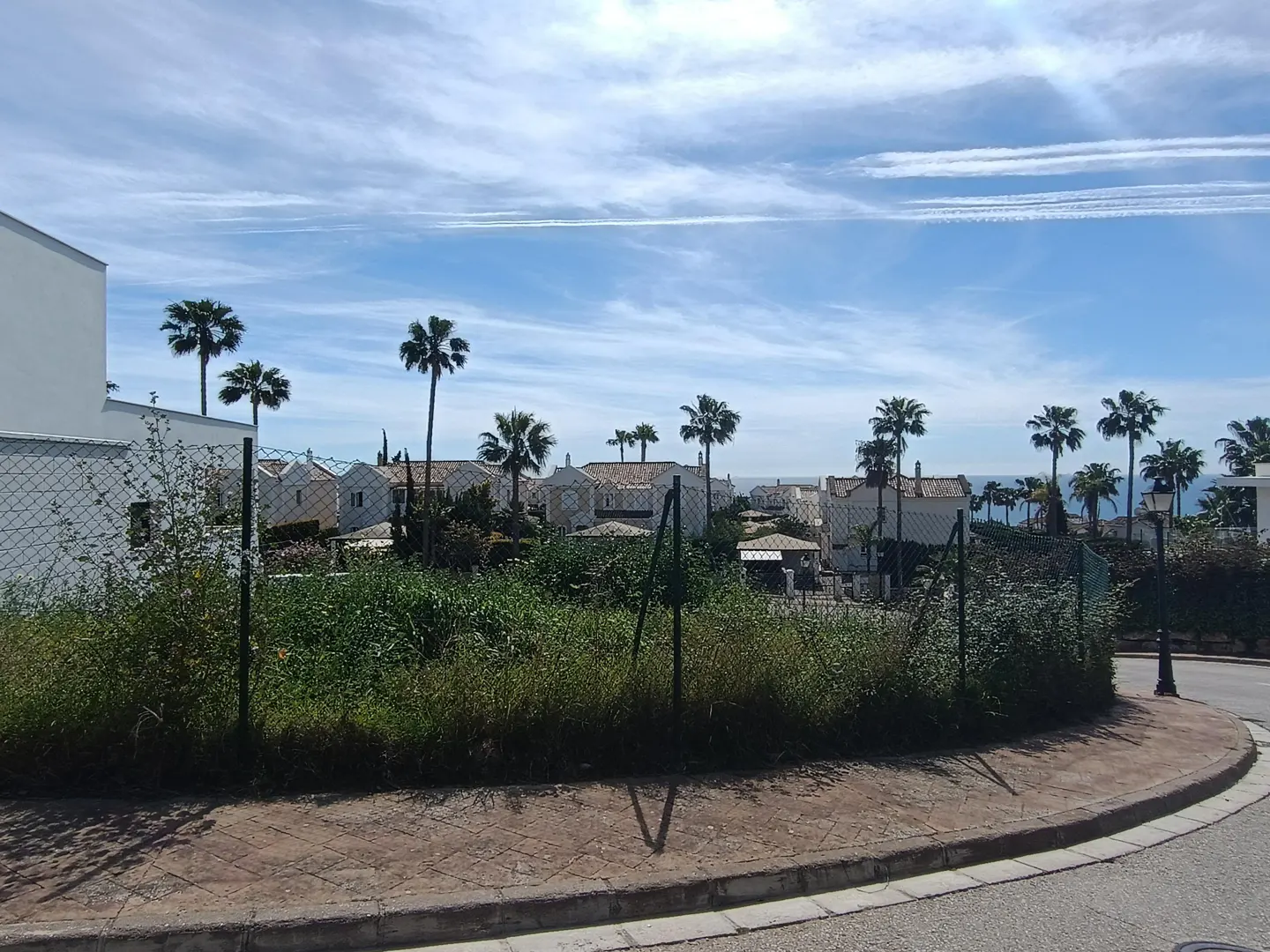 View of a vacant lot with palm trees, houses, and a blue sky with clouds in the background. A chain-link fence surrounds the lot.